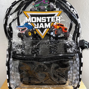 May include: A clear plastic backpack featuring a black and white checkered trim. The front displays a Monster Jam logo with graphics of a purple, a blue shark-themed, and an orange monster truck. It includes a front zippered mesh pocket.