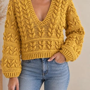 Peut inclure: Un pull à manches longues jaune moutarde avec un décolleté en V profond et un motif texturé en points de noisette sur le corps et les manches, porté avec un jean bleu clair. Le pull présente des poignets et un ourlet côtelés.