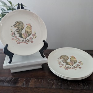 May include: Off-white ceramic plates with a rooster design. The rooster is depicted in shades of green, yellow, and brown, surrounded by floral accents in pink and red. One plate is displayed on a black stand, and three plates are stacked.