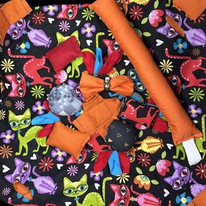 May include: A black fabric cat-themed activity mat with various textures and shapes for cats to play with. The mat features a variety of colorful cats, flowers, and hearts. It includes a long orange pillow, a small red pillow, a gray and white checkered pillow, a blue and red bow, and several small orange pillows.
