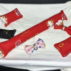 Peut inclure: Un ensemble de jouets en tissu rouge et noir avec l'embl&egrave;me de la maison Gryffondor et les mots "Gryffondor" et "Aastic". Les jouets sont en forme de coussins et d'un long coussin fin. Il y a aussi un n&oelig;ud rose avec un motif blanc et bleu.