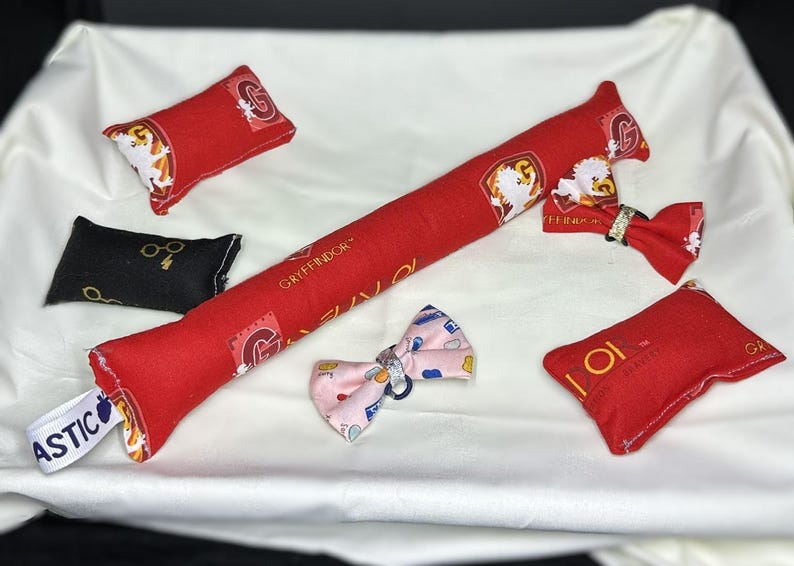 Peut inclure: Un ensemble de jouets en tissu rouge et noir avec l'embl&egrave;me de la maison Gryffondor et les mots "Gryffondor" et "Aastic". Les jouets sont en forme de coussins et d'un long coussin fin. Il y a aussi un n&oelig;ud rose avec un motif blanc et bleu.