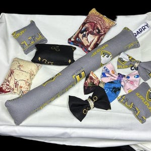 Peut inclure: Une collection d'articles en tissu sur le th&egrave;me d'Harry Potter, comprenant des coussins, une baguette magique et des n&oelig;uds papillon. Les coussins pr&eacute;sentent diff&eacute;rents blasons et motifs de maisons, notamment Gryffondor, Poufsouffle et Serpentard. La baguette est grise avec des accents dor&eacute;s et les n&oelig;uds papillon sont de diff&eacute;rentes couleurs et motifs.
