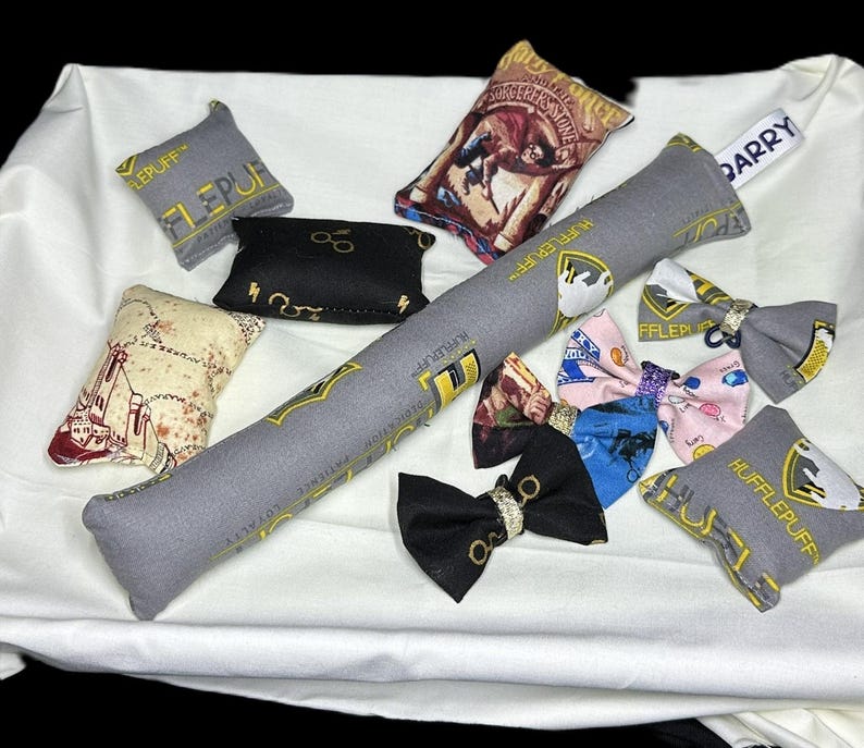 Peut inclure: Une collection d'articles en tissu sur le th&egrave;me d'Harry Potter, comprenant des coussins, une baguette magique et des n&oelig;uds papillon. Les coussins pr&eacute;sentent diff&eacute;rents blasons et motifs de maisons, notamment Gryffondor, Poufsouffle et Serpentard. La baguette est grise avec des accents dor&eacute;s et les n&oelig;uds papillon sont de diff&eacute;rentes couleurs et motifs.