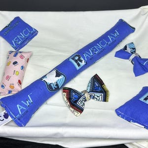 Peut inclure: Un ensemble de pochettes en tissu bleu et rose avec les mots "Ravenclaw", "Wit Learning Wisdom" et "Law Wisdom" imprim&eacute;s dessus. Les pochettes sont d&eacute;cor&eacute;es d'un oiseau, d'un blason et d'autres motifs. Il y a aussi deux n&oelig;uds papillon en tissu bleu avec un motif de blason.