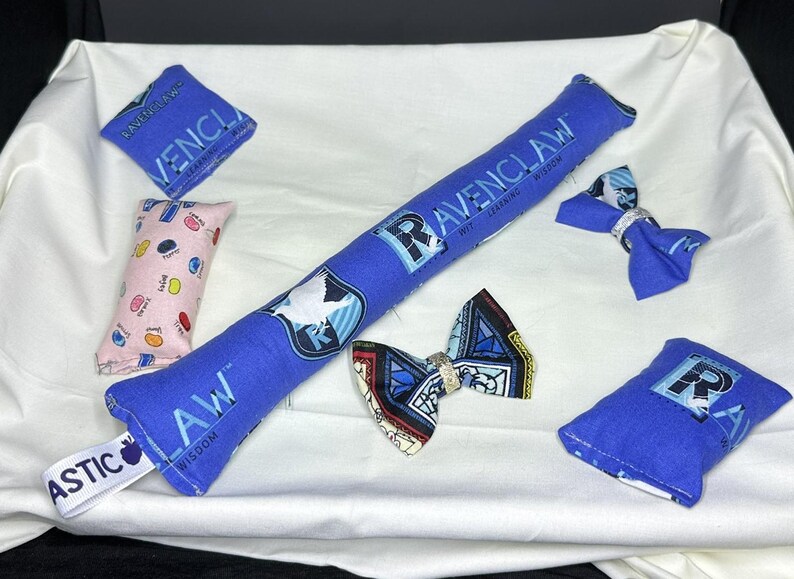Peut inclure: Un ensemble de pochettes en tissu bleu et rose avec les mots "Ravenclaw", "Wit Learning Wisdom" et "Law Wisdom" imprim&eacute;s dessus. Les pochettes sont d&eacute;cor&eacute;es d'un oiseau, d'un blason et d'autres motifs. Il y a aussi deux n&oelig;uds papillon en tissu bleu avec un motif de blason.