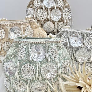 Puede incluir: Cuatro bolsos de noche con cuentas en colores champán, plata y verde mar. Cada bolso presenta adornos florales hechos de lentejuelas y cuentas, con detalles de perlas colgantes. Los bolsos tienen asas de perlas.