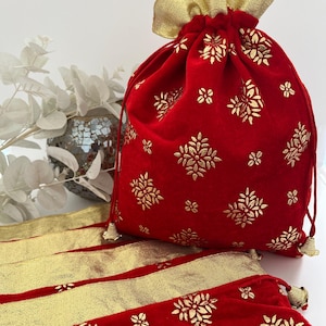 May include: A red velvet drawstring bag with gold floral patterns and a gold ruffled top. The bag is accompanied by matching red fabric with gold trim and floral designs. The bag has a tassel on the drawstring.