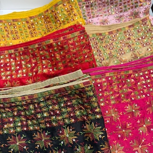 Op de afbeelding: Een verzameling kleurrijke, geborduurde stoffen. De stoffen hebben ingewikkelde patronen met pailletten en bloemmotieven. Kleuren zijn onder meer geel, rood, roze, zwart en groen. De stoffen zijn opgevouwen en tonen het gedetailleerde vakmanschap.