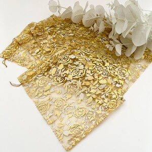Peut inclure: Sacs en tissu transparent de couleur or avec un motif de roses. Les sacs sont faits d'un matériau délicat et sont décorés d'un motif floral répétitif. Une branche d'eucalyptus blanc est en arrière-plan.