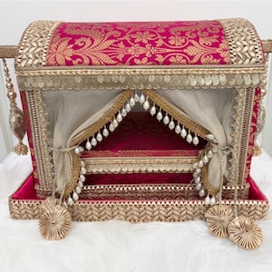 Peut inclure: Un coffre décoratif avec un motif floral fuchsia et or. Il est orné de dentelle dorée, de paillettes et de perles pendantes. L'intérieur présente un tissu drapé et une base rouge foncé.