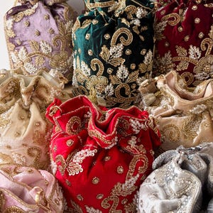 May include: A collection of velvet drawstring bags in various colors, including red, green, and pink. Each bag is adorned with intricate gold embroidery and beadwork, creating a luxurious and ornate appearance. The bags are gathered at the top with a drawstring closure.
