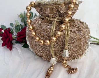 Könnte beinhalten: Ein brauner, bestickter Beutel mit Kordelzug, goldfarbenen Perlen und Quasten. Die Tasche hat aufwendige Blumenmuster und einen Perlenhenkel. Der Hintergrund umfasst eine rote Rose und weißen Stoff.