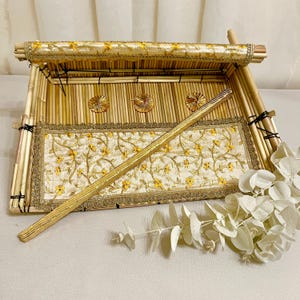 Op de afbeelding: Een decoratief bamboe dienblad met een gouden geborduurde stoffen voering en een houten stok. Het dienblad heeft een deksel en is versierd met gouden accenten.