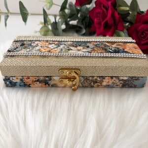 May include: A rectangular decorative box with a gold clasp. The box features a floral pattern in shades of blue, orange, and cream. The top and sides are embellished with gold glitter and a row of clear rhinestones. The box is resting on a white, fluffy surface.