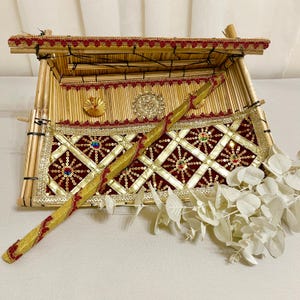 May include: A decorative bamboo box with a red and gold patterned fabric lining. The box is open and a gold stick with red accents is resting on top of the fabric. White flowers are in the foreground.