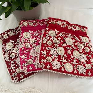 May include: Three embroidered drawstring bags in shades of red and pink. Each bag features intricate floral patterns in white and gold thread, with a pearl bead trim along the bottom edge. The bags are likely for jewelry or small gifts.