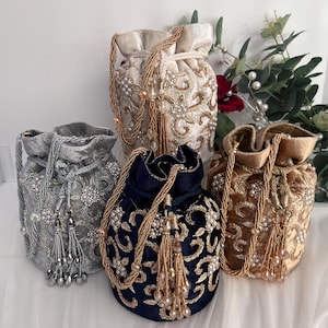 May include: Four velvet drawstring bags in silver, navy, gold, and cream. Each bag features intricate embroidery, beaded accents, and tassel details. The bags have braided drawstrings and are displayed on a white surface.