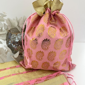 May include: A pink drawstring bag featuring a gold paisley pattern and gold trim. The bag is secured with a pink drawstring and has a gold fabric top. Matching pink fabric with gold trim is in the foreground.