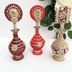 May include: Three decorative red and gold perfume bottles with intricate designs and embellishments. The bottles have round tops and are adorned with gold thread, beads, and sequins.
