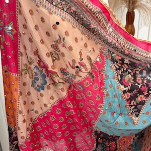 Könnte beinhalten: Ein lebendiger, gemusterter Schal mit Patchwork-Design. Der Schal zeigt eine Mischung aus floralen und geometrischen Mustern in Rosa, Orange, Blau und Schwarz. Verziert mit kleinen, reflektierenden Pailletten, hat er einen leuchtend rosa Rand.