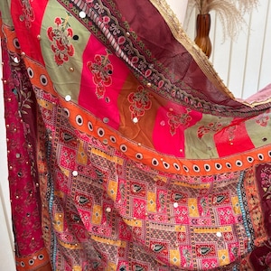 Könnte beinhalten: Ein farbenfrohes Textil mit einem Patchwork-Design. Der Stoff weist eine Mischung aus roten, rosa, orangefarbenen und grünen Farbtönen auf, verziert mit floralen Mustern und kleinen, reflektierenden Verzierungen. Ein goldener Besatz säumt den Rand.