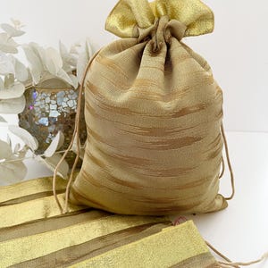 Peut inclure: Un sac cadeau doré avec cordon de serrage et un tissu doré assorti. Le sac est fait d'un tissu texturé avec des rayures horizontales. Le cordon de serrage est de couleur marron clair. Le sac est noué en haut.