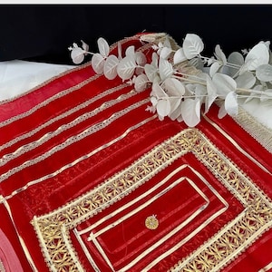 May include: A bright red sheer fabric with gold trim and decorative patterns. The fabric features gold ribbon and a central rectangular design. White floral accents are placed on the fabric.
