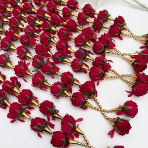 May include: Several red rose garlands with gold-coloured accents. Each rose is paired with a gold-toned bell-shaped piece and strung together with gold beads. The roses have green leaves at the base.