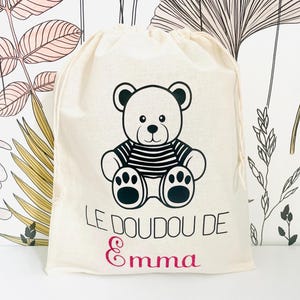 May include: A beige drawstring bag featuring a black and white cartoon bear wearing a striped shirt. The text "LE DOUDOU DE Emma" is printed on the bag in black and pink glitter. The bag is made of a soft fabric.