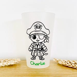 Puede incluir: Un vaso esmerilado con un diseño de pirata de dibujos animados en blanco y negro. El pirata lleva un sombrero con una calavera, un parche en el ojo y una espada. El nombre "Charlie" está impreso en verde en la parte inferior.