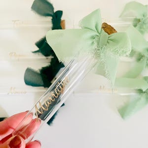 May include: A clear glass tube with a cork stopper and a light green ribbon bow. The tube has the word "Marraine" in gold script. Other tubes with names and dark green ribbons are in the background. The theme is likely for a special event.