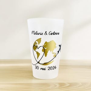 Peut inclure: Un verre givré avec un motif de mappemonde doré, un cœur et un avion noir. Le texte "Mélanie & Antoine" et "30 mai 2026" sont imprimés sur le verre. Le verre est cylindrique et posé sur une surface claire.