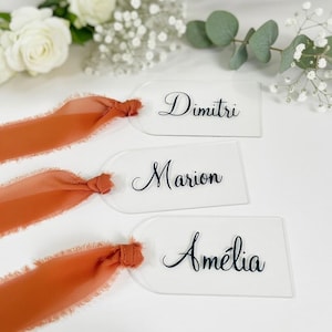 Può includere: Segnaposto in acrilico trasparente con top arrotondati, con nomi in elegante scrittura nera: Dimitri, Marion e Amélia. Ogni carta è legata con un nastro arancione bruciato. Fiori bianchi e verde sullo sfondo.