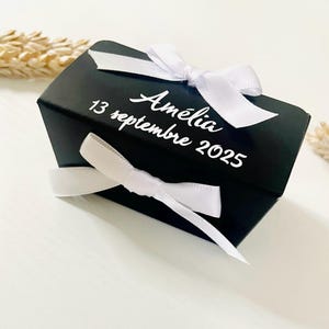 May include: A black gift box with white text and a white ribbon tied in a bow. The text on the box reads "Amélia 13 septembre 2025". The box is rectangular and has a satin ribbon wrapped around it.