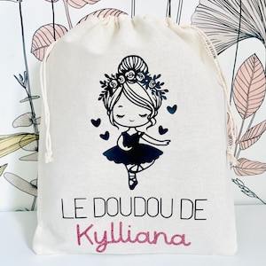 May include: A cream-colored drawstring bag featuring a black illustration of a ballerina with a floral crown and hearts. The text "LE DOUDOU DE Kylliana" is printed on the bag in black and pink glitter.