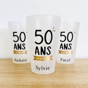 Op de afbeelding: Drie matte plastic bekers met zwarte tekst "50 ANS" en de zin "ça pique" op een gouden banner. De namen "Nathalie", "Sylvie" en "Farid" staan onder elke beker afgedrukt.