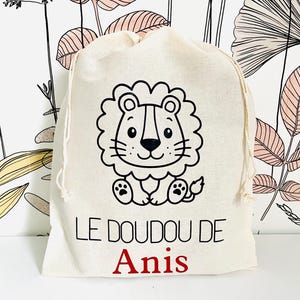May include: A beige drawstring bag featuring a black and white cartoon lion illustration. The text "LE DOUDOU DE Anis" is printed on the bag in black and red. The bag is made of a soft, natural fabric.