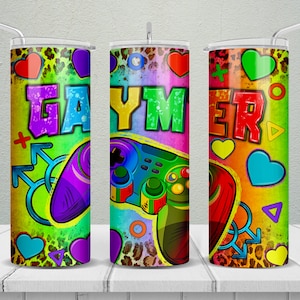May include: A colorful tumbler featuring the word "GAYMER" in graffiti-style lettering. The design includes game controllers, hearts, and other gaming-related symbols. The background has a leopard print pattern and a rainbow gradient.