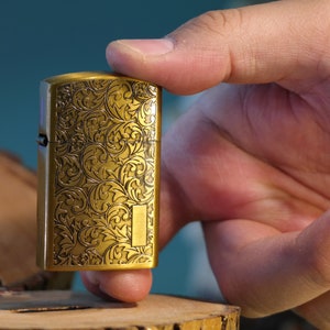 Puede incluir: Un encendedor dorado con un intrincado patrón floral grabado en la superficie. El encendedor está siendo sostenido por una mano.