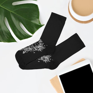 Puede incluir: Calcetines negros con un diseño gráfico blanco de un pájaro y la palabra "FELICIS". Los calcetines son de material suave y adecuados para el uso diario. Se encuentran sobre una superficie blanca.
