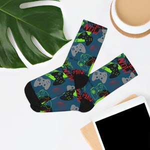 May include: A pair of blue and black socks with a pattern of green and gray video game controllers, red and green text that says "Now Play" and "Pin", and the words "Ha-Ha" in red.