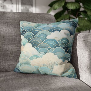 May include: Square decorative pillow with a watercolor design featuring blue and white cloud-like shapes with gold accents. The pillow is placed on a gray sofa, adding a touch of elegance to the room's decor.