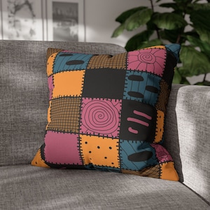 Könnte beinhalten: Quadratisches Kissen mit Patchwork-Design. Das Kissen hat Quadrate in Orange, Schwarz, Türkis und Pink mit gestickten Details und skurrilen Strichzeichnungen. Das Kissen liegt auf einem grauen Sofa.