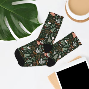 May include: A pair of dark green socks featuring a repeating pattern of birds, trees, and floral elements in white, red, and brown. The socks have black heels and toes, and are displayed on a white surface.
