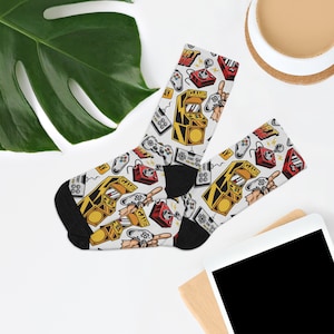 May include: A pair of white socks with a colorful retro video game design. The socks feature images of joysticks, game controllers, and arcade cabinets. The text "Start Game" is printed on the socks.