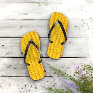 Puede incluir: Un par de chanclas con un diseño de mazorca de maíz amarillo brillante. Las sandalias tienen correas negras y están sobre una superficie de madera blanca desgastada. La imagen evoca un ambiente veraniego y juguetón.