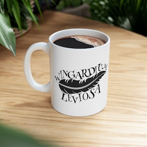 Puede incluir: Taza de cerámica blanca llena de café oscuro. La taza presenta el texto "Wingardium Leviosa" con un gráfico de pluma negra. La taza está sobre una superficie de madera clara.