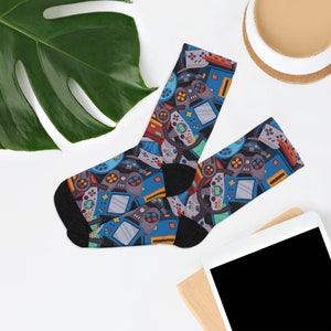 May include: A pair of black socks with a colorful pattern of video game controllers, consoles, and other gaming-related graphics. The socks have a black heel and toe, and the design includes blue, gray, red, and yellow elements.