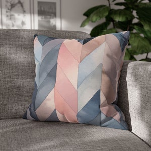 Pode incluir: Almofada decorativa quadrada com um padrão de chevron em aquarela em tons de azul, rosa e cinza. A almofada está colocada em um sofá cinza, criando uma estética suave e convidativa.